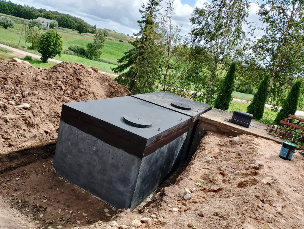 Duży, czarny betonowy zbiornik podziemny (np. szambo lub deszczówka) z włazami, świeżo wkopany w ziemię na terenie prywatnej posesji.