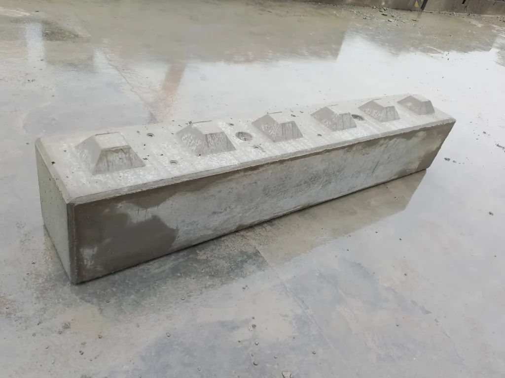 Długi, prostokątny betonowy blok typu lego z pięcioma wypukłymi elementami na górze, leżący na mokrej posadzce.