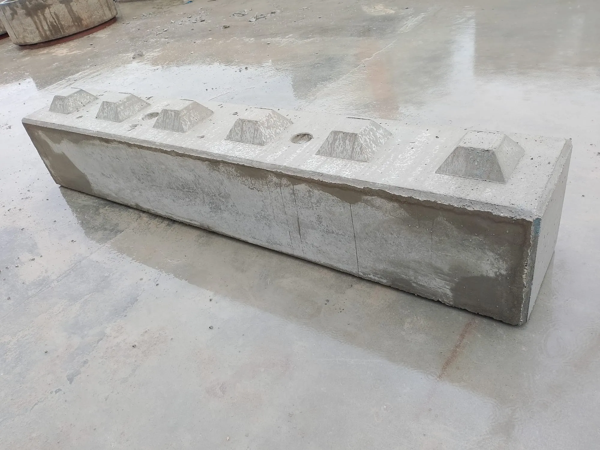 Długi, prostokątny betonowy blok typu lego z pięcioma wypukłymi elementami na górze, leżący na mokrej posadzce.