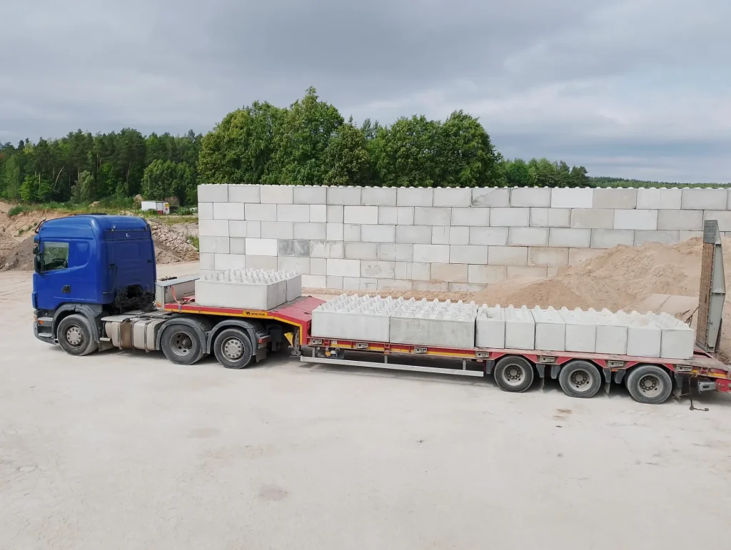 Niebieska ciężarówka Scania z naczepą przewożąca betonowe bloki oporowe na tle muru ze stabilizujących bloków betonowych.